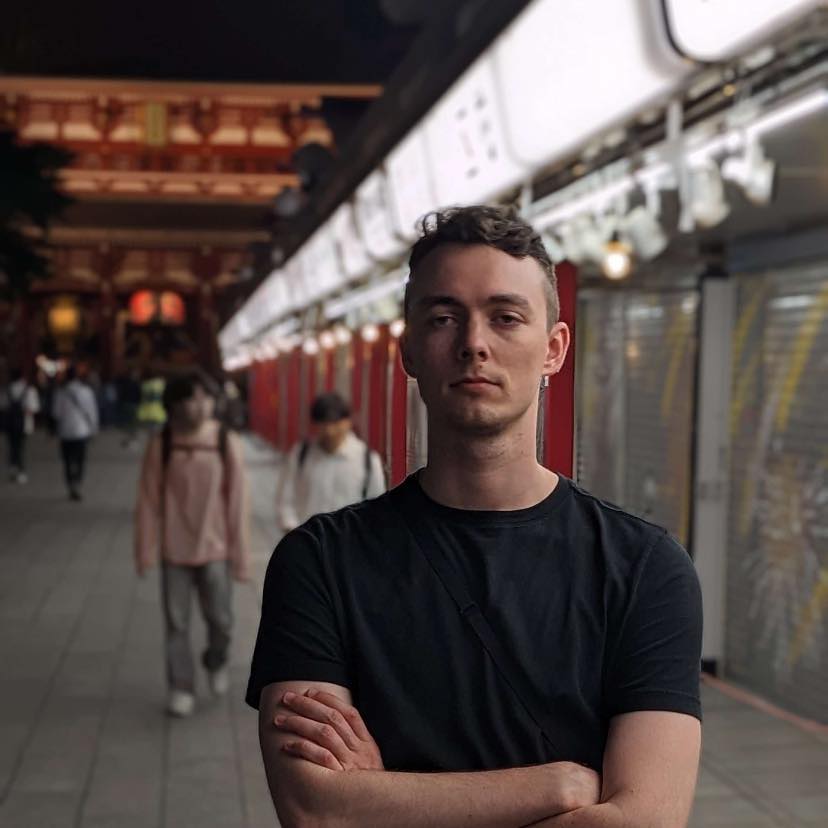 A headshot of Forrest standing in front of a temple in Tokyo. Dramatic lighting bathes one side of his face in even white light. He's wearing a plain black t-shirt. It's a little self-serious.